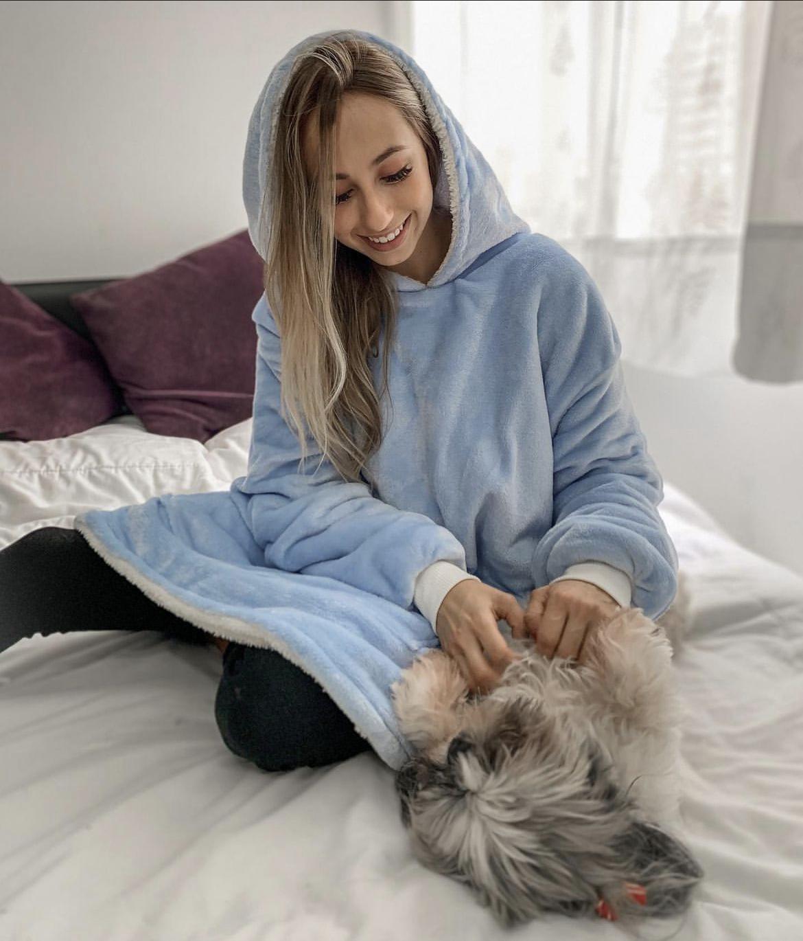 Mujer riendo sentada en la cama usando un saco cobija azul claro jugando con una perrita