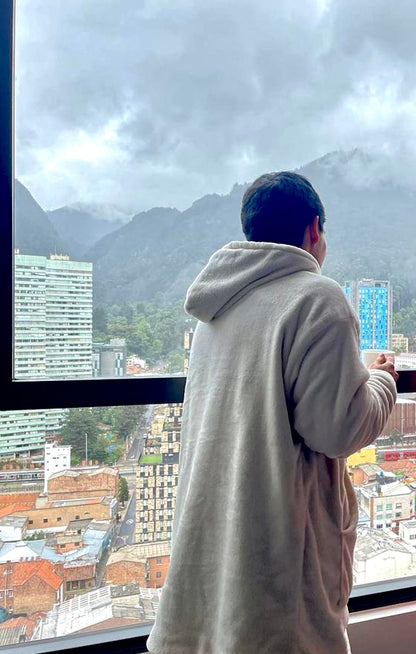 Hombre usando un saco cobija gris claro parado de espaldas, mirando por la ventana mientras se toma un café