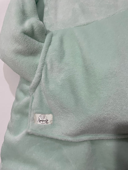 Bolsillo de un saco cobija color verde menta, con las mangas dentro del bolsillo y una marquilla beige satinada de la marca Apapacho Mio