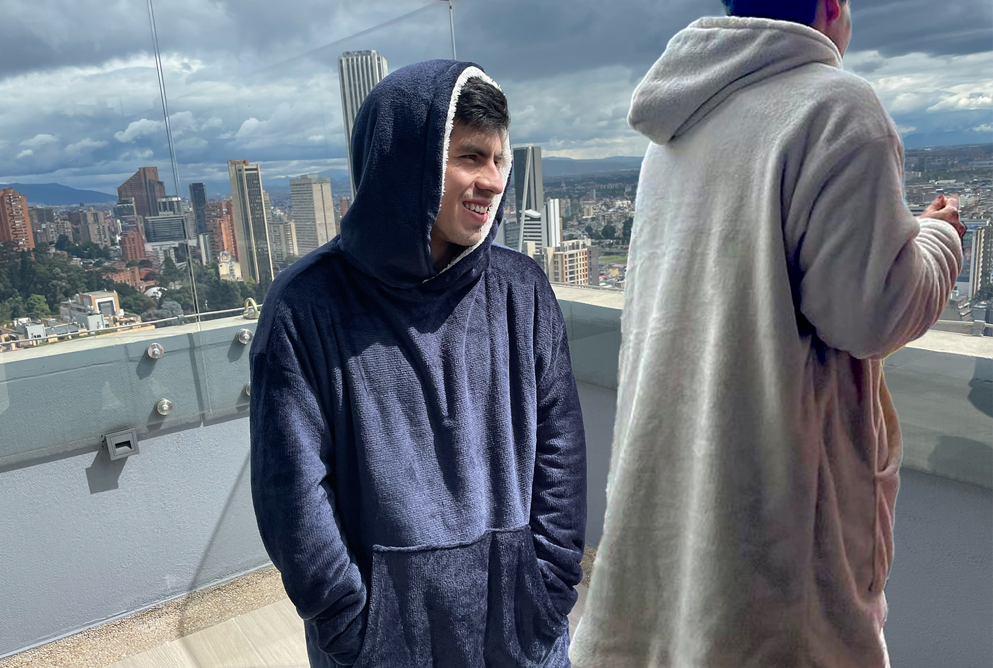 Hombre sonriendo usando un saco cobija doble faz por el lado azul oscuro y hombre de espaldas parado a su lado de espaldas mirando a la ciudad desde una terraza y usando el saco gigante doble faz por el lado gris claro 